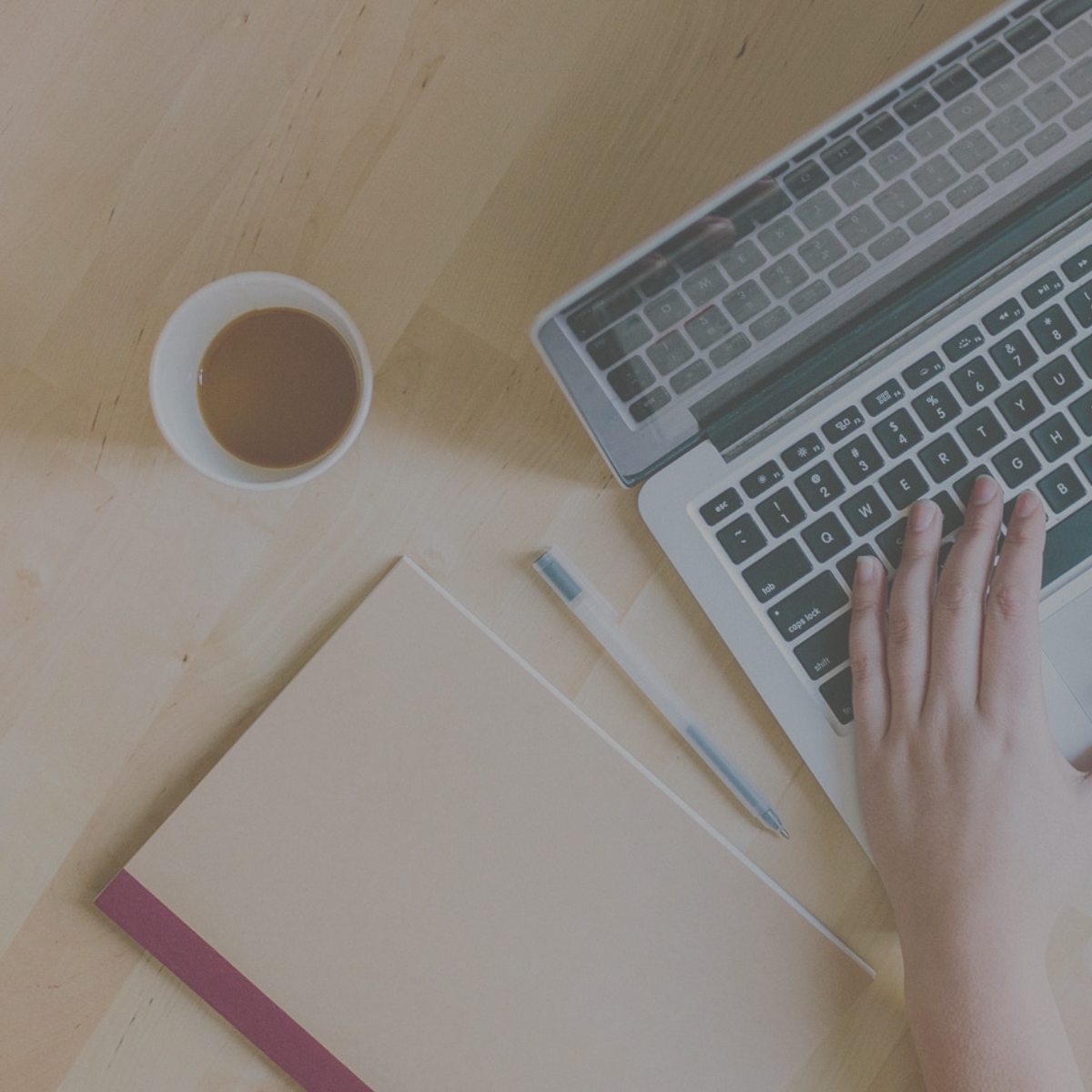 A person typing on a laptop with a journal, pen and mug aesthetically beside them.